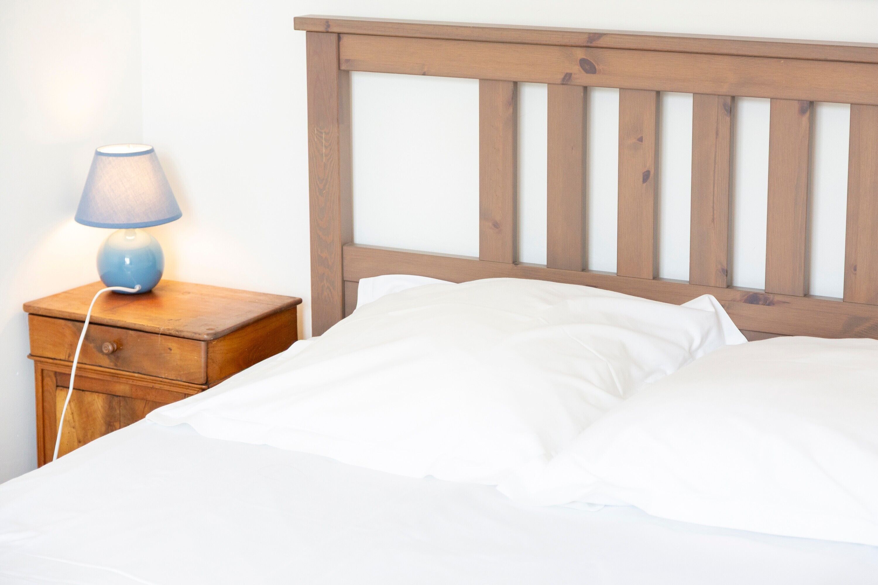 A bed with white linens, a wooden headboard, and a bedside table with a lamp.