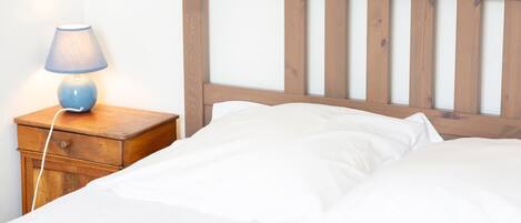 A bed with white linens, a wooden headboard, and a bedside table with a lamp.