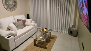 A modern living room with a white sofa, a glass coffee table, and a large TV on the wall.