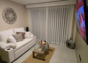 A modern living room with a white sofa, a glass coffee table, and a large TV on the wall. - New condo, 2.8 km airport SJO, 3 bedrooms, pool, A/C, WiFi, parking (Río Segundo)