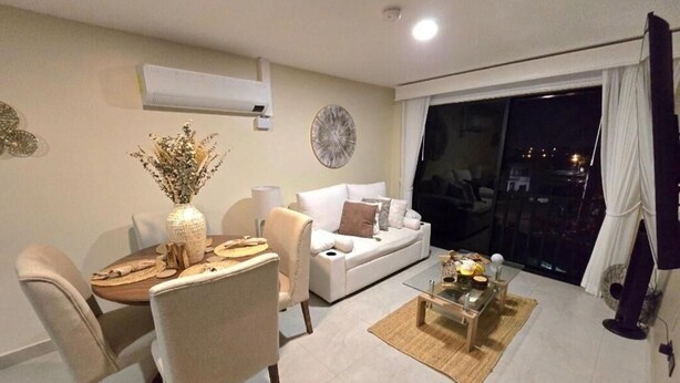 A modern living room with a dining area, a white sofa, a coffee table, and a large window with a view of the city at night.