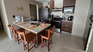 A modern kitchen with a granite countertop, wooden chairs, a stainless steel refrigerator, and a microwave above the stove.