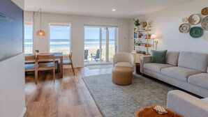 Uma sala de estar ampla com uma grande janela com vista para a praia, um espaço de estar acolhedor e uma estante com objetos decorativos.