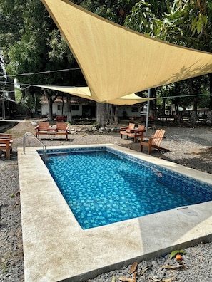 A swimming pool with a tiled edge, surrounded by a concrete deck. There are wooden chairs and a shaded area with a yellow canopy.