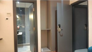 A hotel room with a glass door leading to a bathroom, a dark-colored door, and a small shelf. - Hotel Golden Leaf (Ranchi)