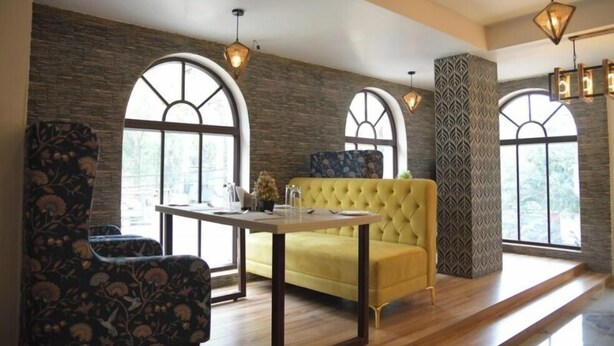 A room with a yellow sofa, a table, and a patterned armchair. There are large windows with arched tops and a brick wall. - Hotel Golden Leaf (Ranchi)