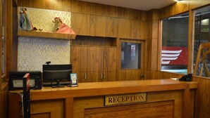 A reception area with wooden paneling, a reception desk labeled "RECEPTION," a computer, and a window with a view of a red building. - Hotel Lalbagh Gardenia (Bengaluru)