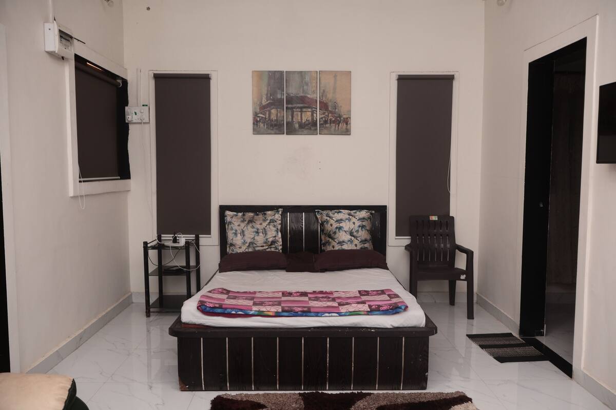 A bedroom with a bed, a painting above it, a chair, and a desk.