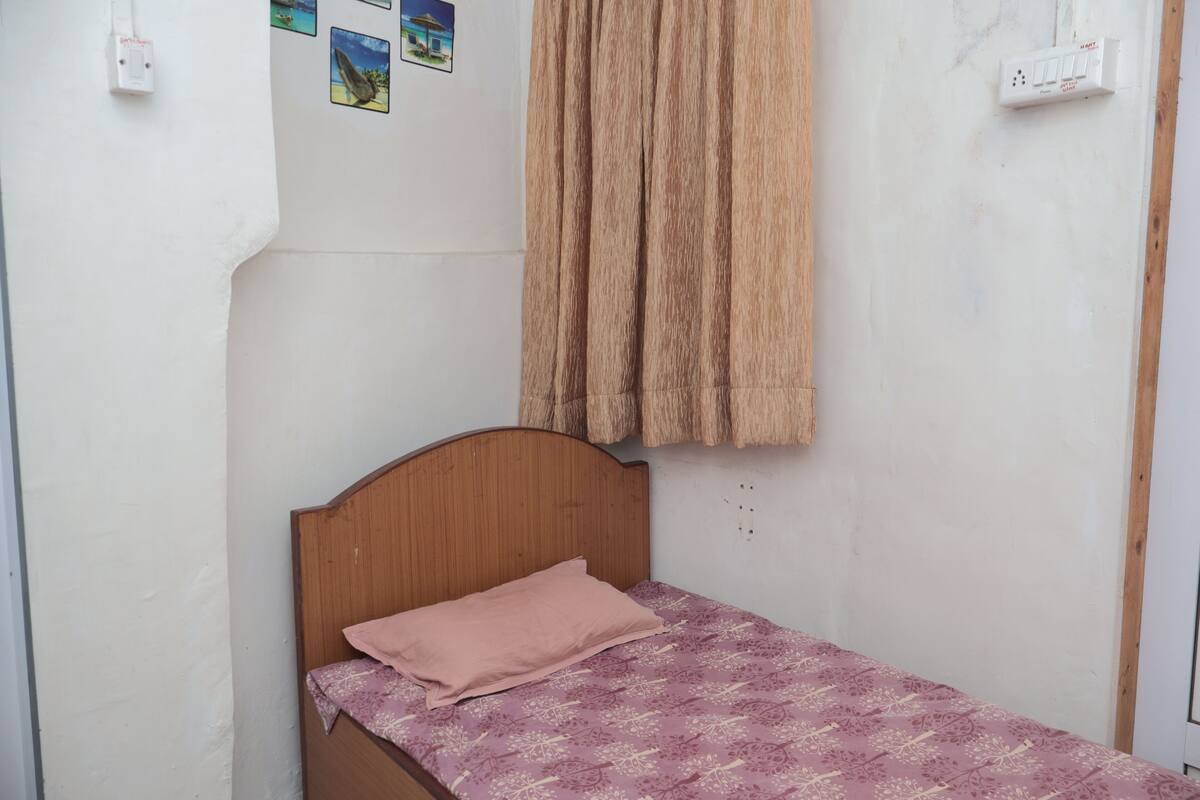 A small, single-bed room with a wooden headboard, a pink pillow, and a patterned bedspread. There are two electrical outlets on the wall and a curtain hanging above the bed.