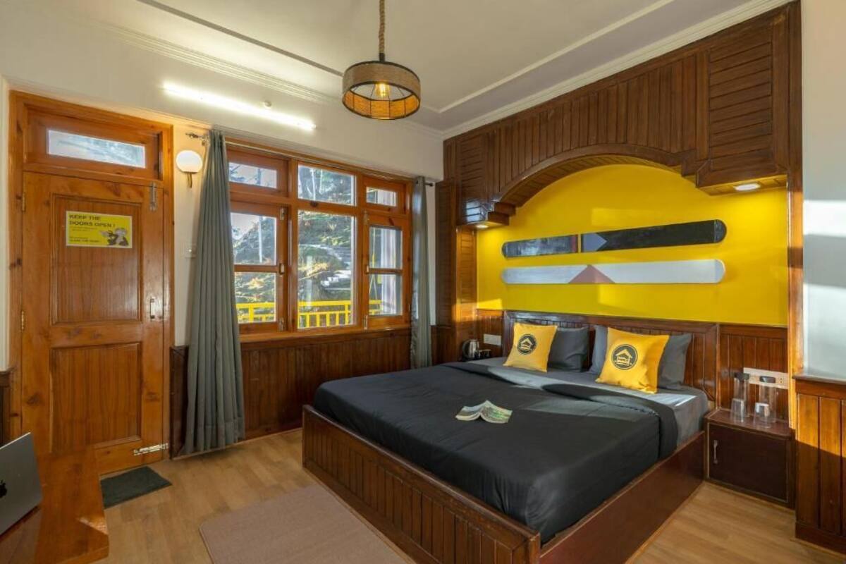 A hotel room with a large bed, wooden paneling, a yellow accent wall, and a view of the outdoors.