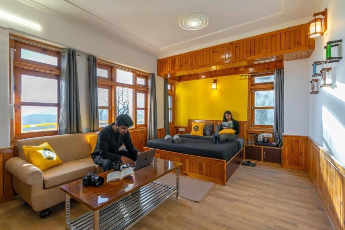A room with a wooden ceiling, a bed with pillows, a sofa with a cushion, a table with a laptop, and a window with curtains.