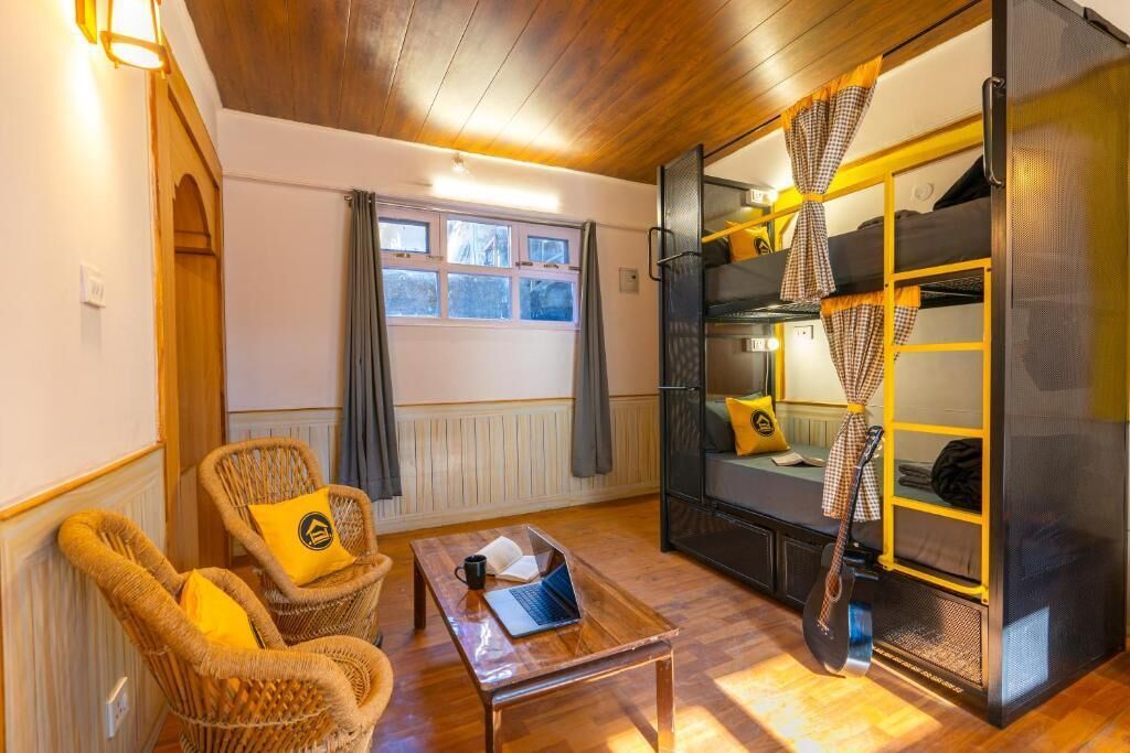 A cozy room with a wooden ceiling, a window with curtains, a sofa with cushions, a wicker chair, a small wooden table with a laptop, and a bunk bed with yellow and black bedding.