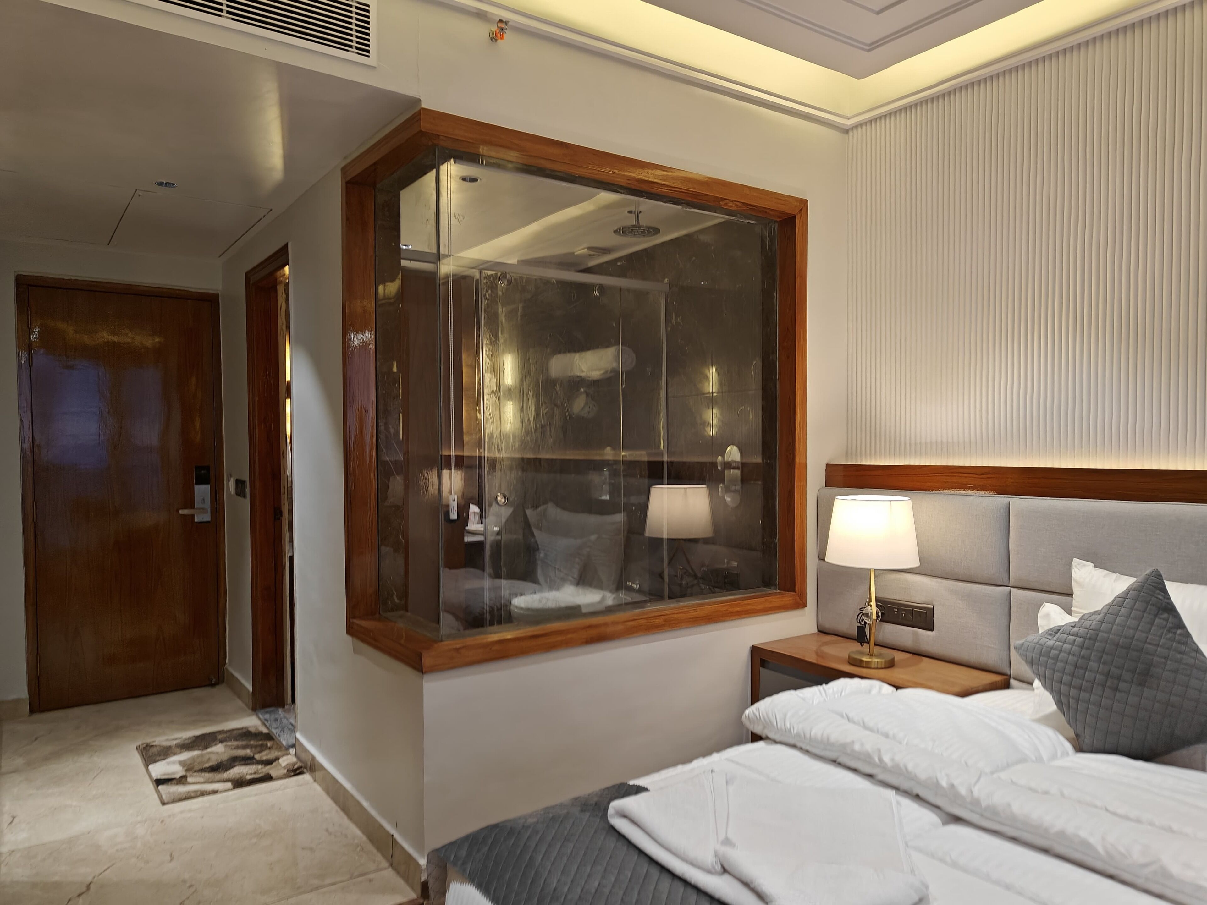 Una habitación de hotel con un ventanal grande, una cama con ropa blanca, una mesita de noche con una lámpara y una puerta de madera.
