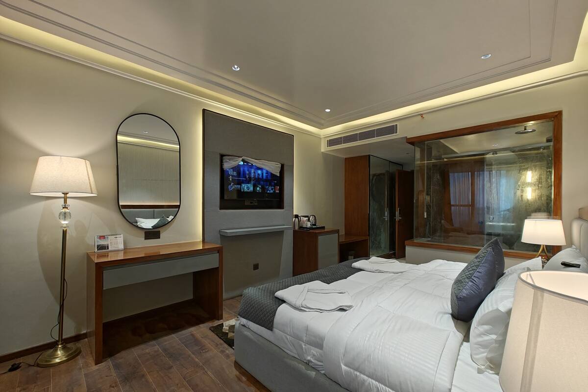 A hotel room with a bed, a desk, a TV, a mirror, and a shower area.
