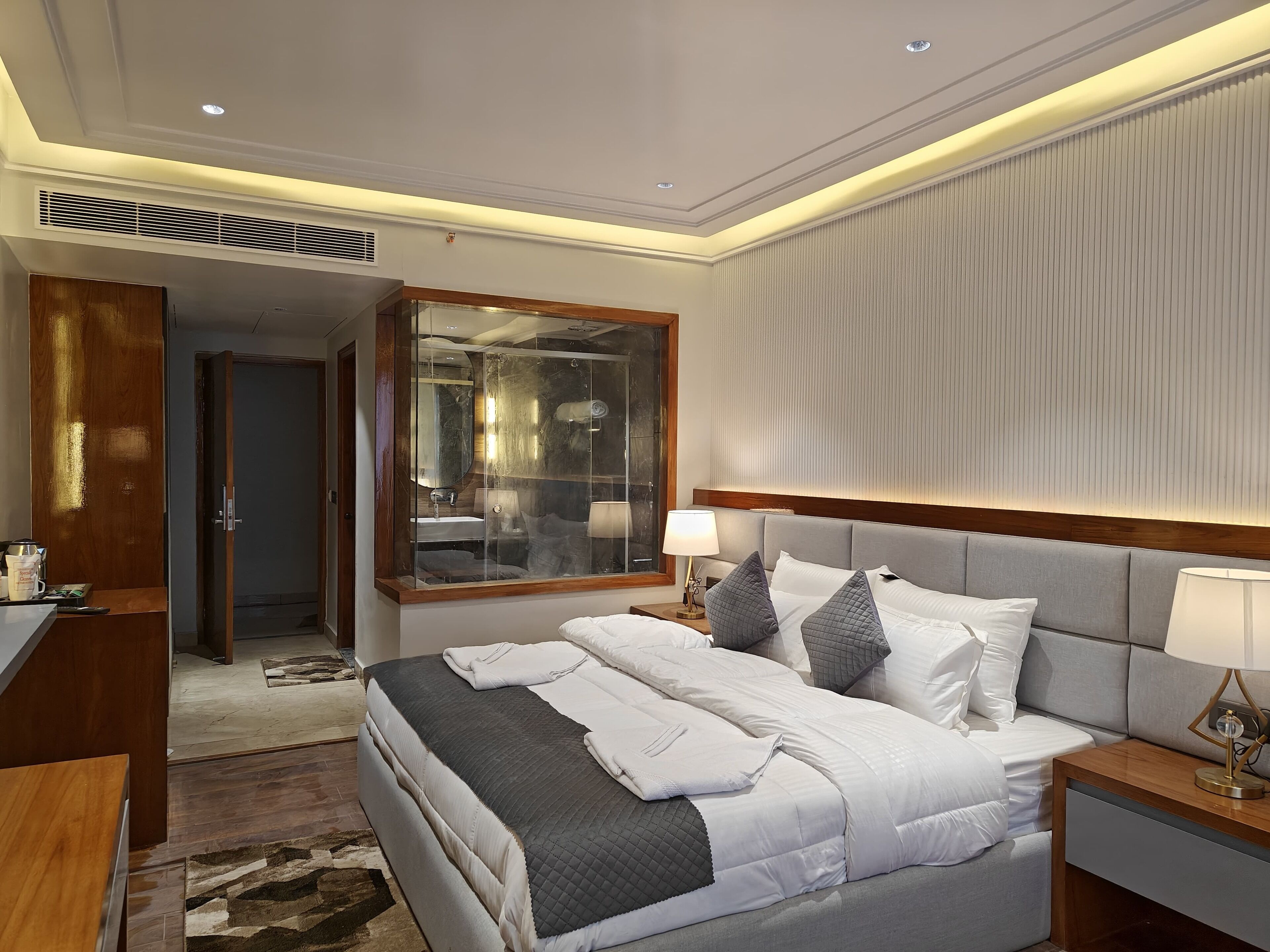 Quarto de hotel moderno com uma cama grande, mesas de cabeceira, um espelho e vista para o banheiro.