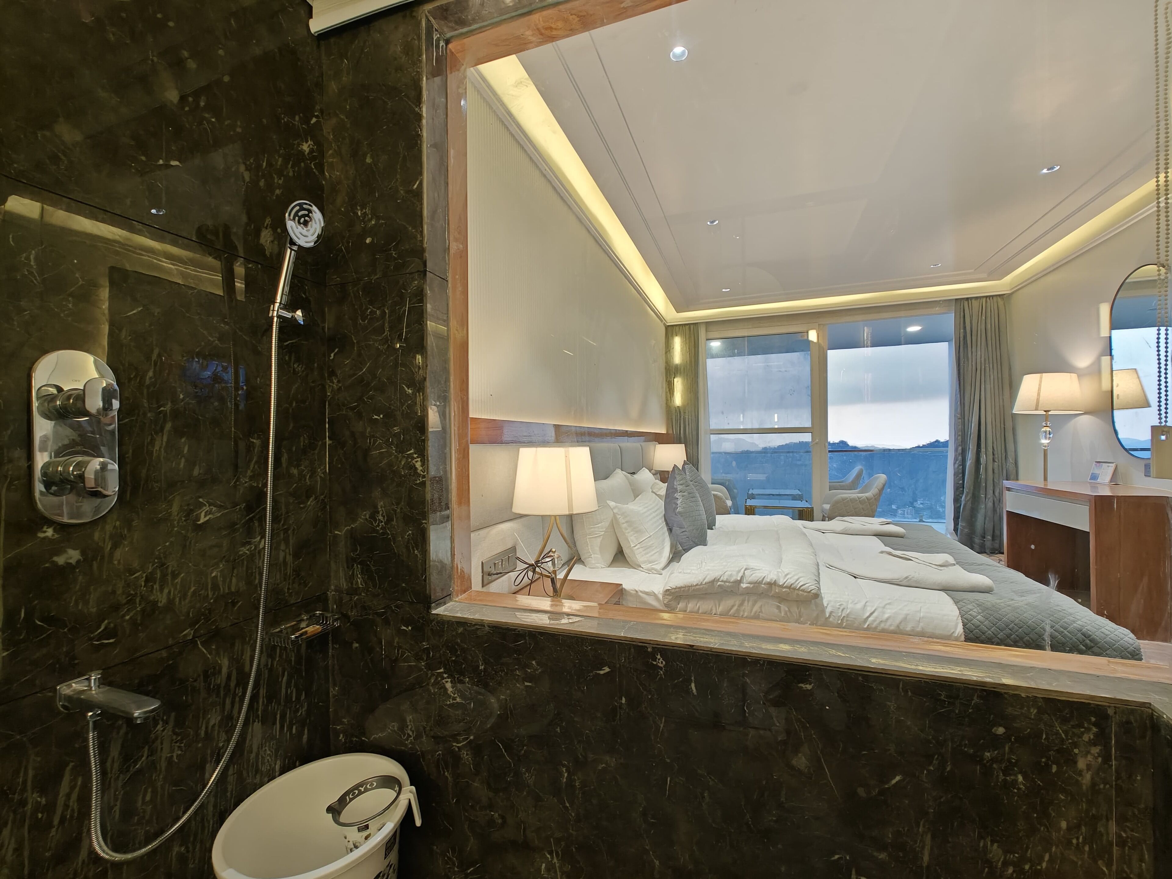 Habitación de hotel moderna con un espejo grande que refleja la cama y el baño, una zona de ducha con cabezal de ducha de mano y vistas a la montaña a través de la ventana.