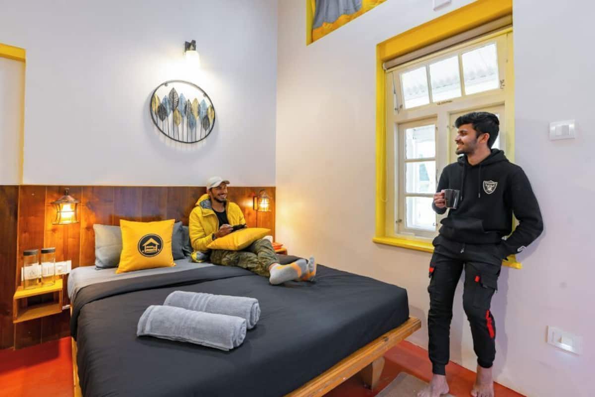 Ein Hotelzimmer mit einem Bett, einem Nachttisch mit Lampe, einem Fenster mit gelben Rahmen und einer Wandleuchte.
