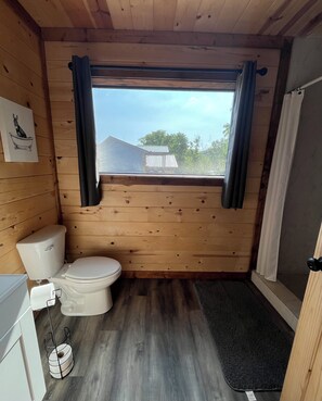 Baño de una pequeña cabaña de madera, con inodoro, ventana con cortinas y vista a una casa exterior.