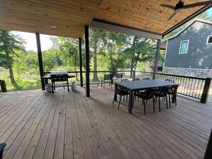 Terraza de madera con mesa, sillas, una parrilla y vistas a una casa y árboles.