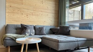Un salon moderne avec un canapé d’angle gris, une petite table ronde et une fenêtre donnant sur une clôture à treillis.