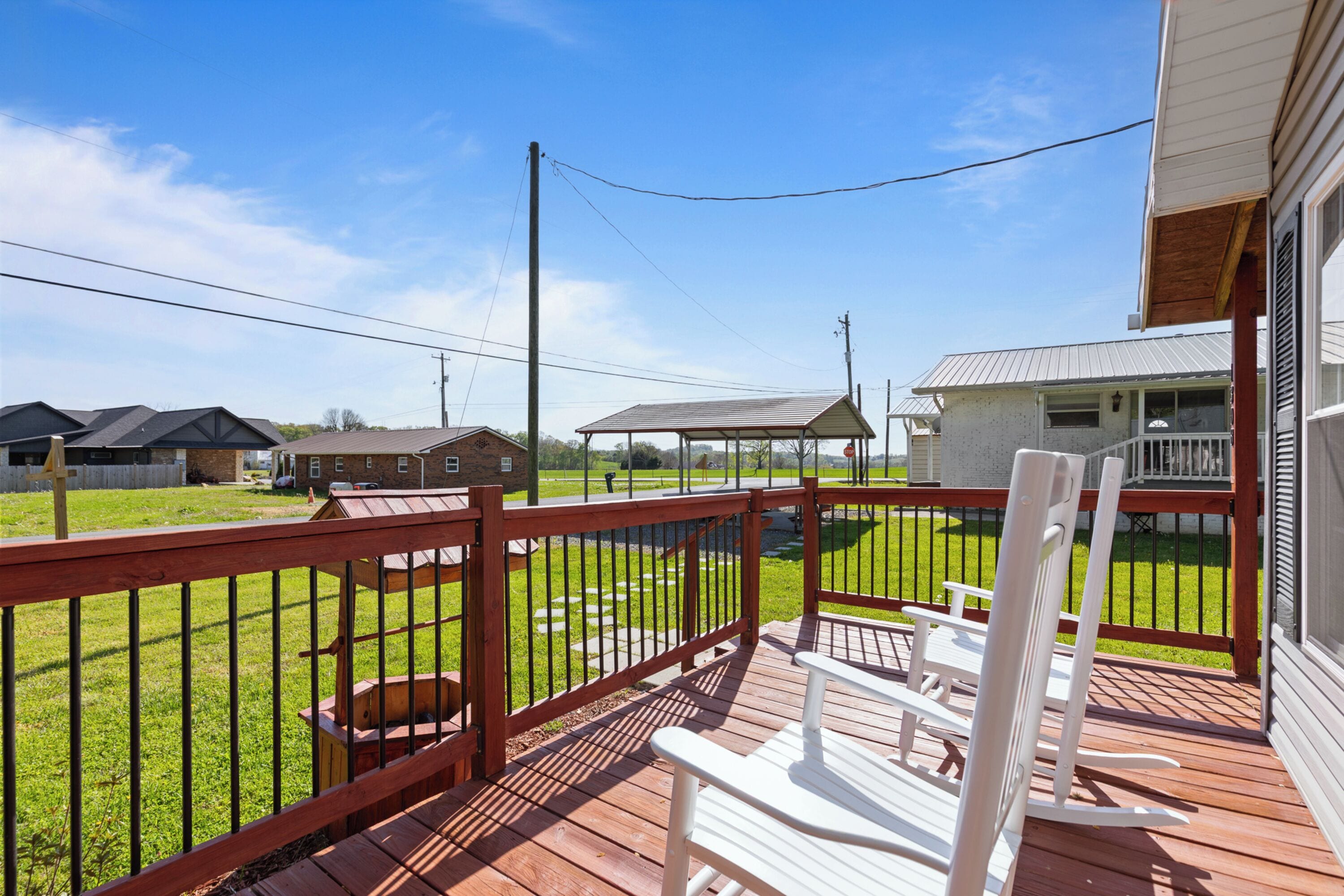 A wooden deck with a railing, white chairs, and a view of a residential area with houses and a clear sky.