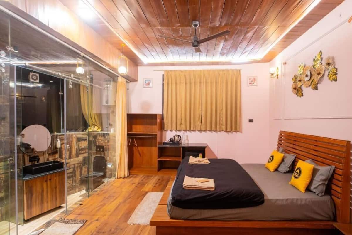 A bedroom with a wooden ceiling, a bed with grey and yellow pillows, a wooden bench, a glass shower enclosure, and a window with curtains.
