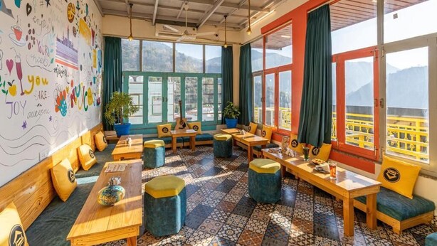 A cozy cafe with wooden tables and blue cushioned seating, large windows with mountain views, and a colorful wall with doodles. - The Hosteller Rishikesh, Upper Tapovan (Uttarakhand)