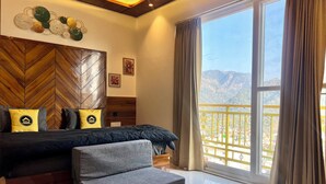 A bedroom with a bed, a grey sofa, a wooden herringbone wall, a large window with curtains, and a view of mountains. - The Hosteller Rishikesh, Upper Tapovan (Uttarakhand)