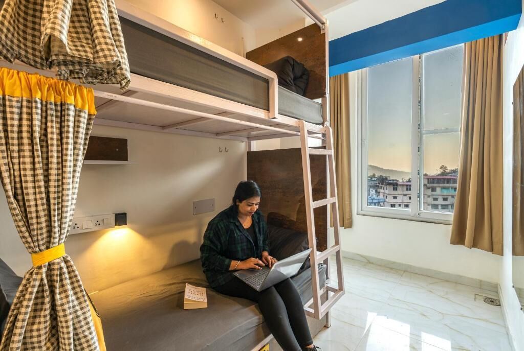 A room with bunk beds, a laptop on a seat, and a window with a view of buildings.