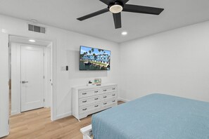 Quarto com cama, televisão na parede, ventilador de teto e cômoda branca.