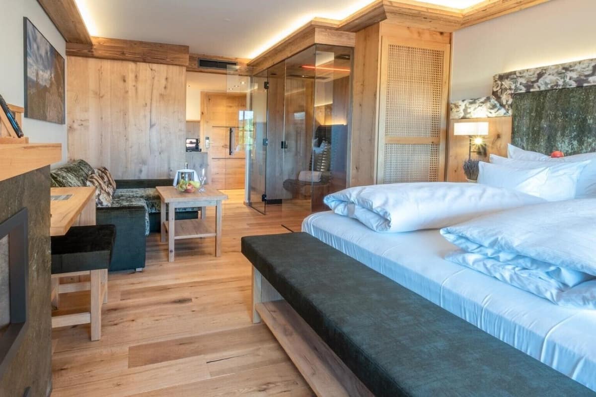 Et moderne hotelværelse med trægulv, seng med hvide sengetøj, et skrivebord med en computer og et opholdsområde med sofa og stole.
