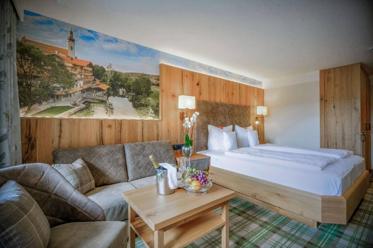 Et hotelværelse med seng, sofa, træsenggavl, et landskabsmaleri og et bord med en flaske og frugt.