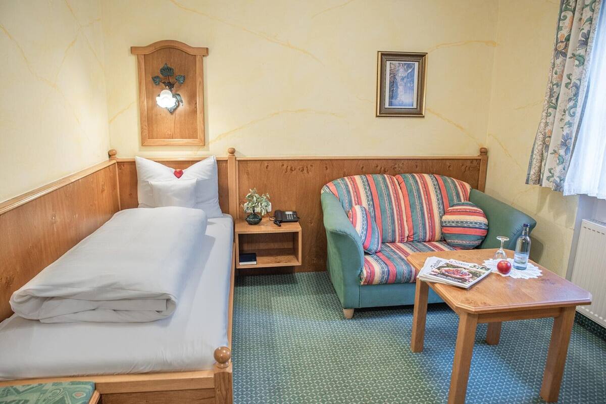 Et hotelværelse med en seng, en grøn sofa, et trænatbord med en telefon og en lille plante, et billede i ramme på væggen og et bord med et magasin og et glas.