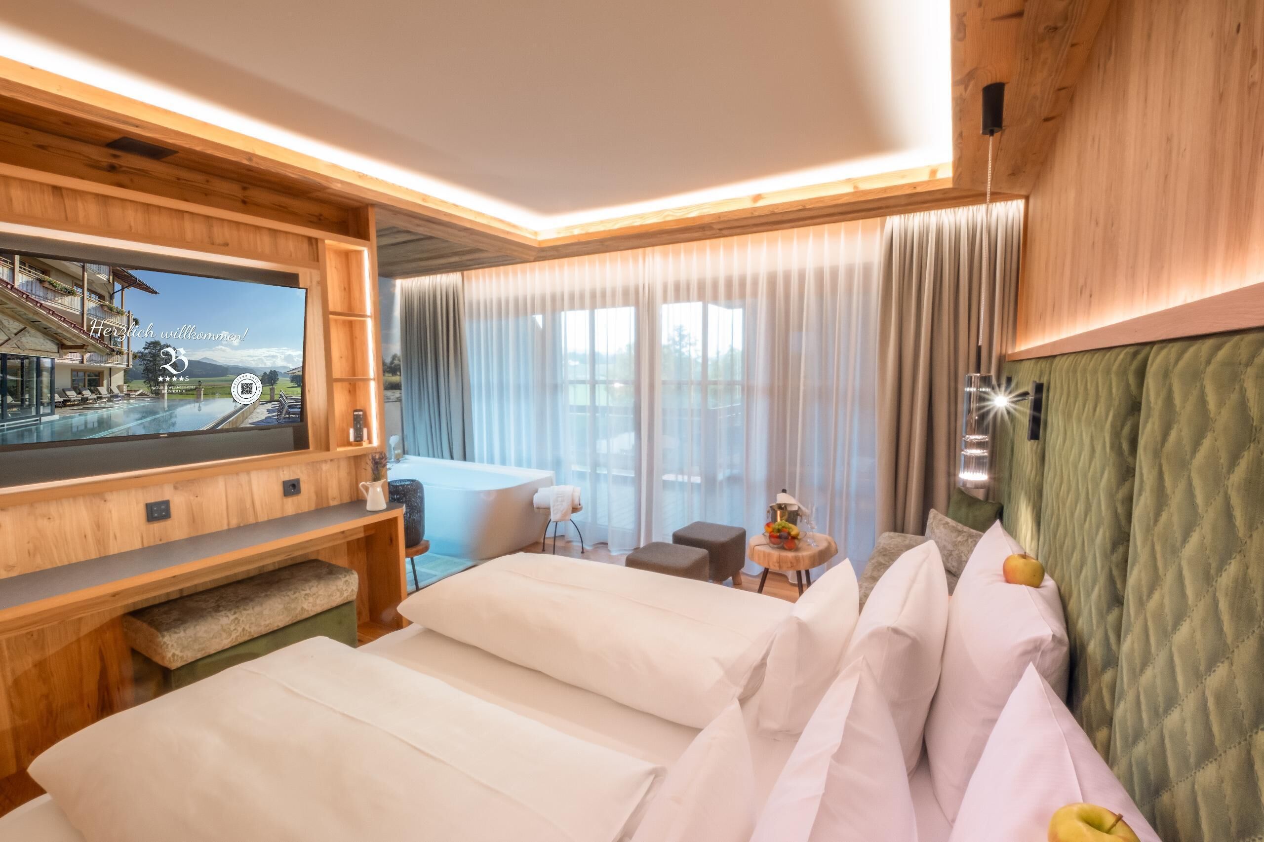 A modern hotel room with a large TV, a bed, a seating area, and a view of a pool and mountains.