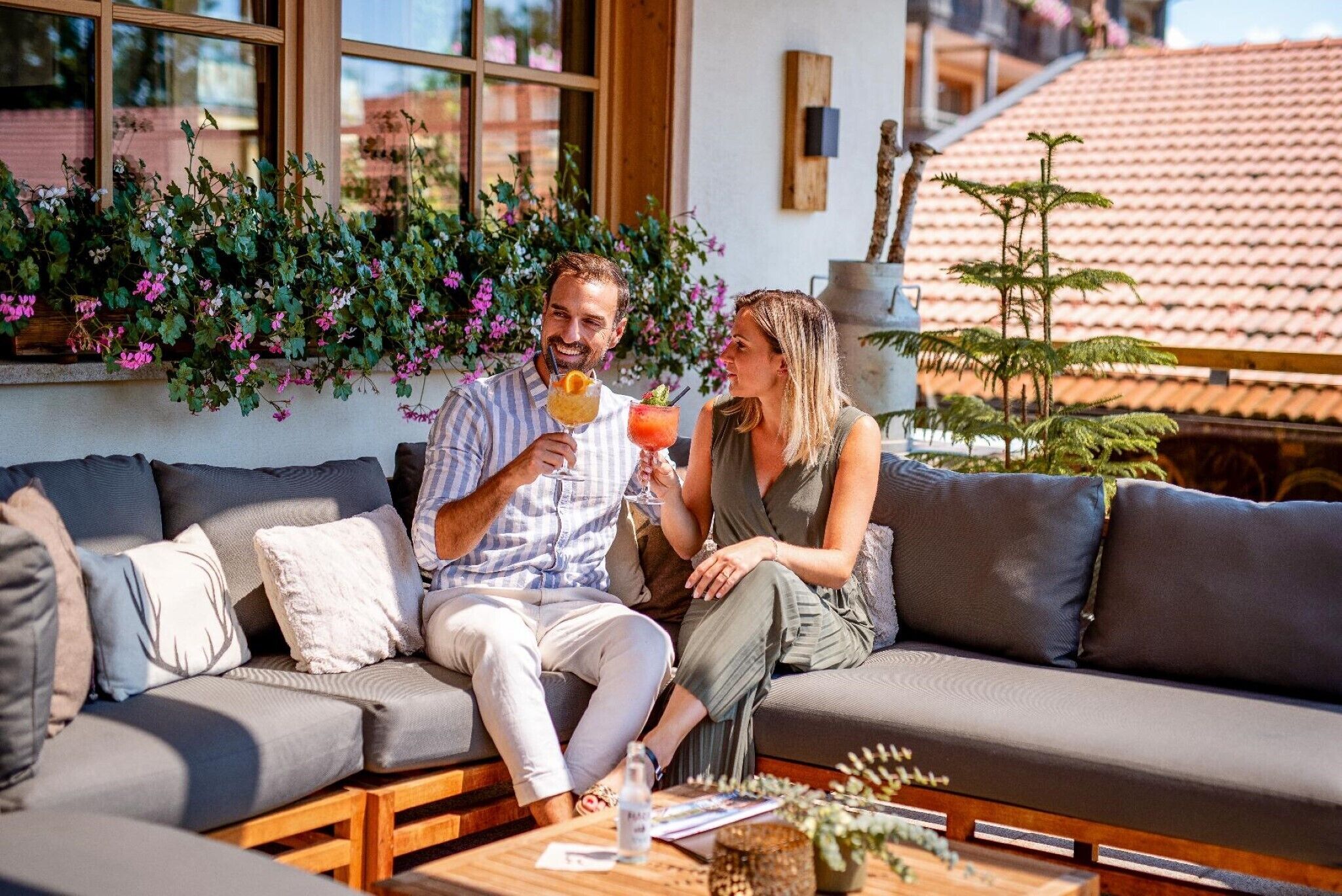 A couple sitting on a gray sofa with a wooden coffee table in front, a window with a view of a red-tiled roof, and a planter with green foliage.