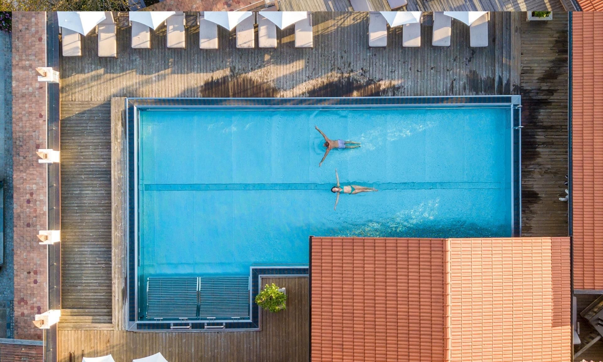 Vista aérea de una piscina rectangular con dos personas nadando, rodeada por una terraza de madera y un muro de ladrillo.