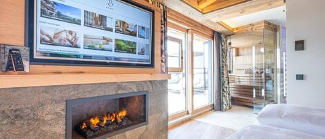 A modern hotel room with a stone fireplace, a large TV screen displaying hotel information, and a view of the outdoor area.