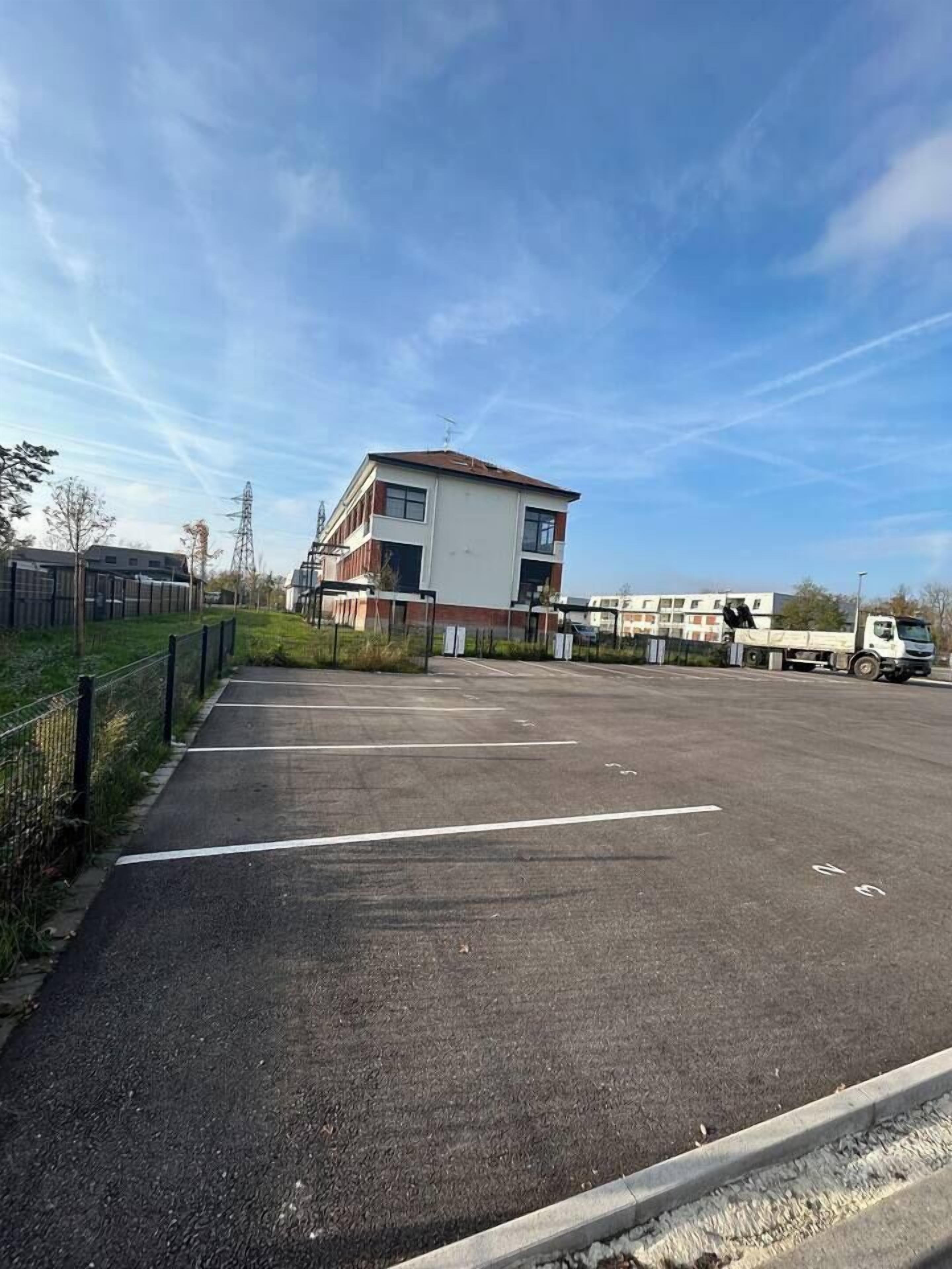 A parking lot with a building in the background, a fence, and a clear sky.