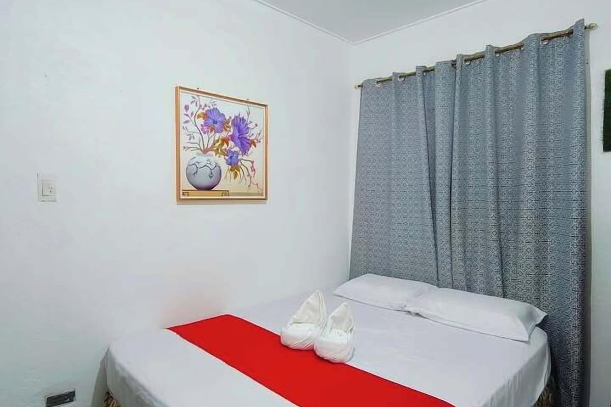 A hotel room with a bed, a red blanket, a pillow, a vase with flowers, and a curtain.