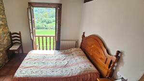 Un dormitorio con pared de piedra, una cama de madera con colcha floral, una silla de madera y vista a un paisaje verde a través de un ventanal abierto.