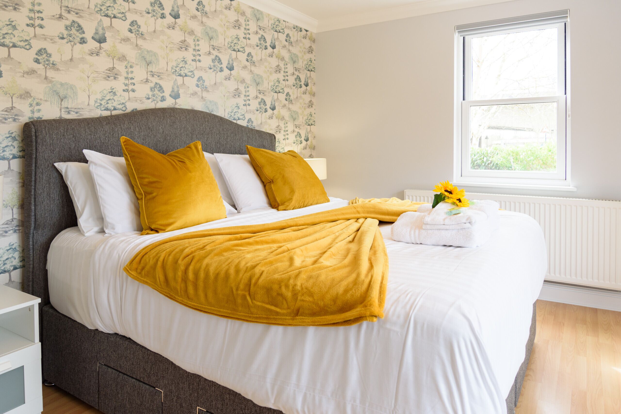 A neatly made bed with white and mustard bedding, a grey headboard, and a window with a view of greenery.