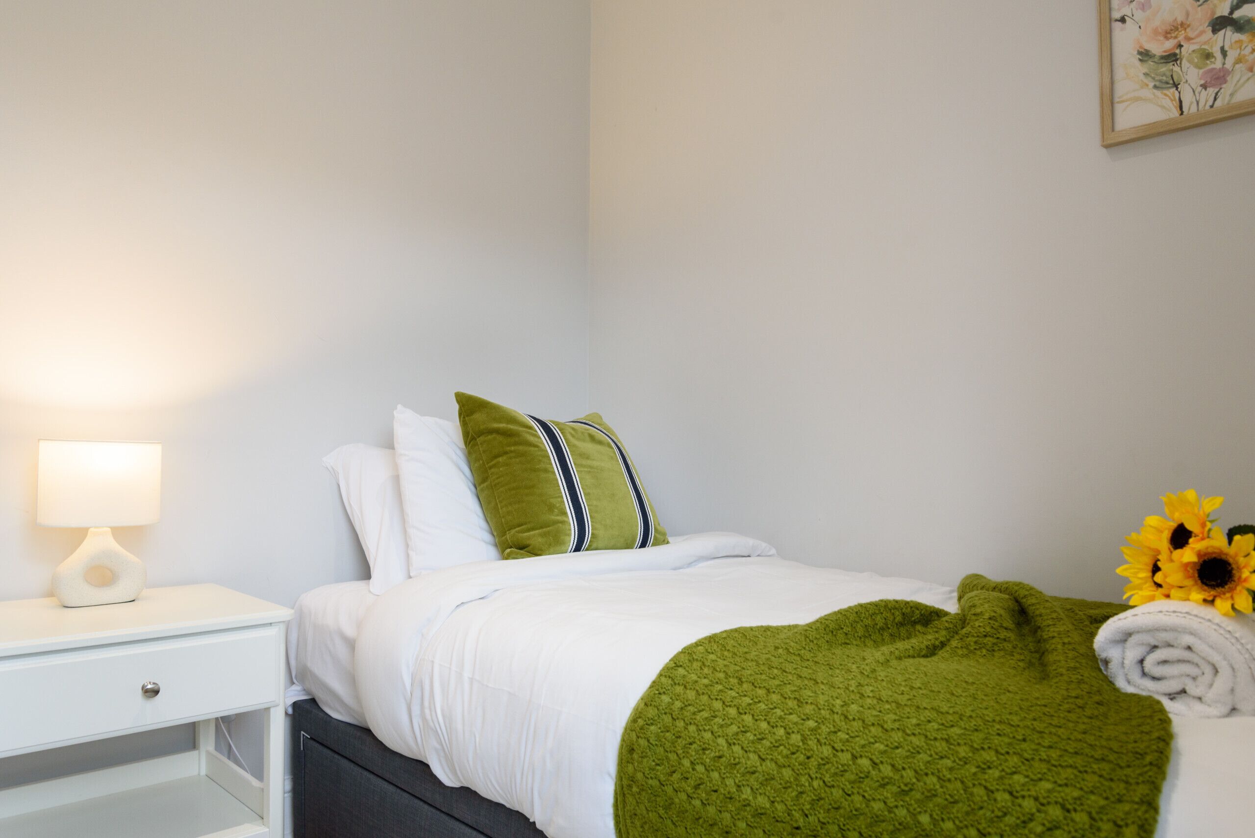 A neatly made bed with white bedding and a green throw blanket, a white bedside table with a lamp, and a wall-mounted painting.