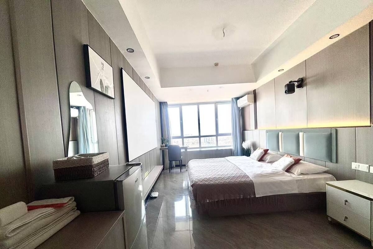 A modern hotel room with a large bed, a desk, and a chair. There is a window providing a view of the city.