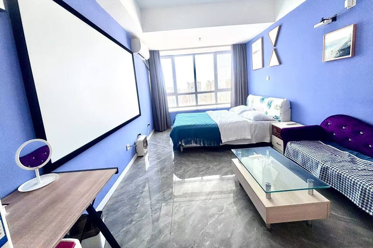 A modern hotel room with a large bed, a sofa, a glass coffee table, and a desk with a chair.