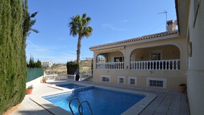 A swimming pool with a white tiled deck, a palm tree, and a building with a balcony.