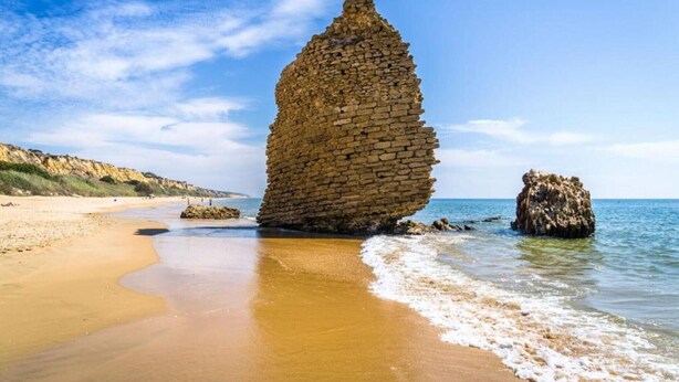 A coastal scene with a prominent stone structure on the beach, waves gently lapping at its base.