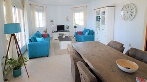 A modern living room with two blue sofas, a wooden dining table, and a TV in the background.