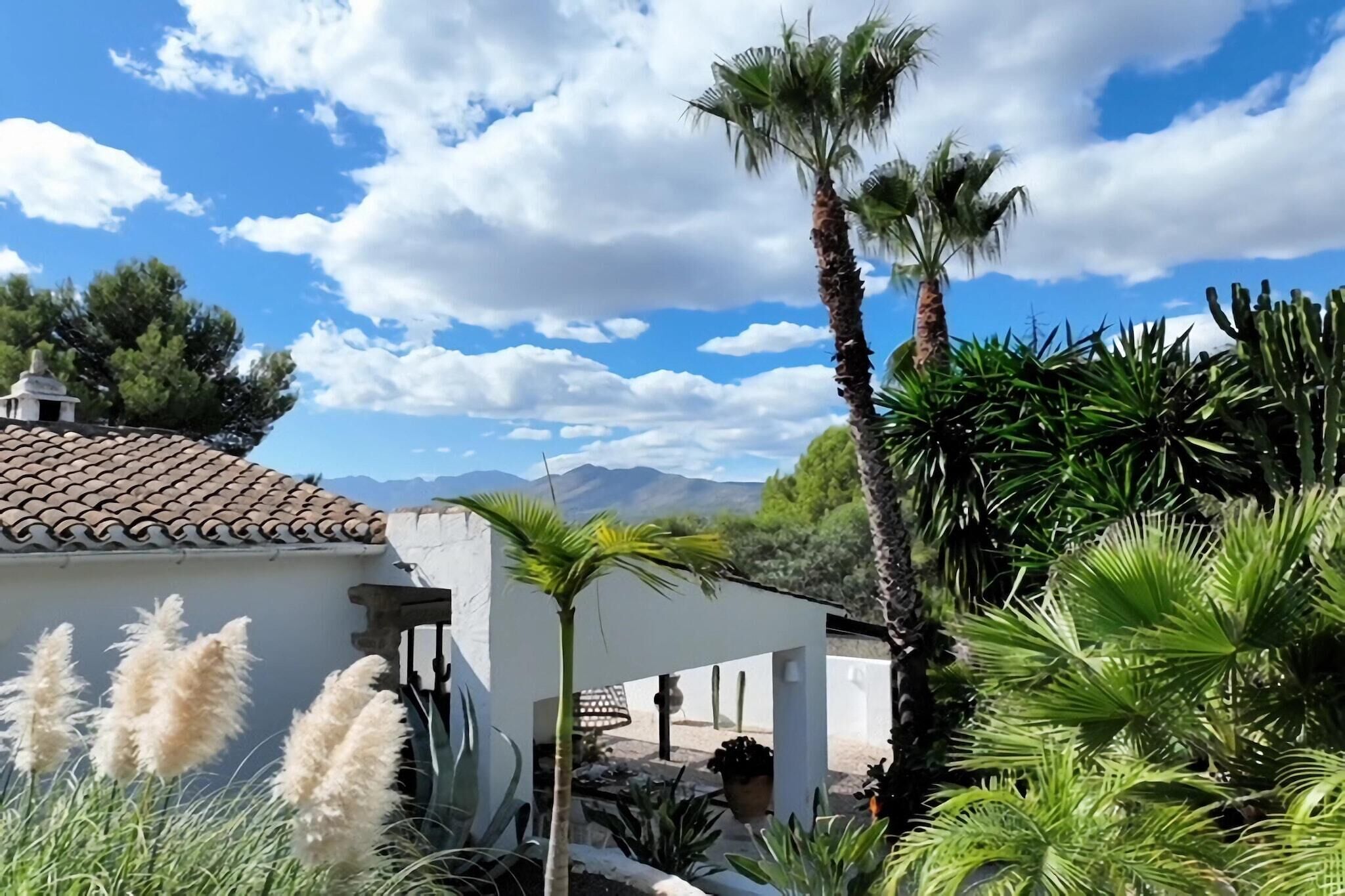 A white building with a tiled roof, surrounded by cacti and pampas grass, with mountains in the background.