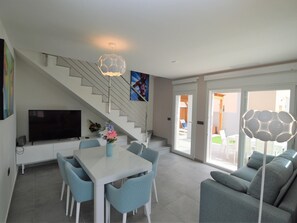 A modern living room with a white staircase, a dining table with chairs, a sofa, and a TV.