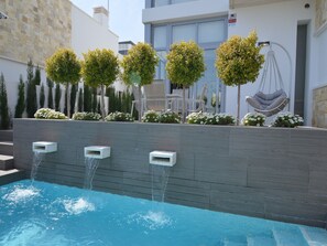 Piscina com três fontes, rodeada por um muro de pedra, com área de estar externa e árvores.